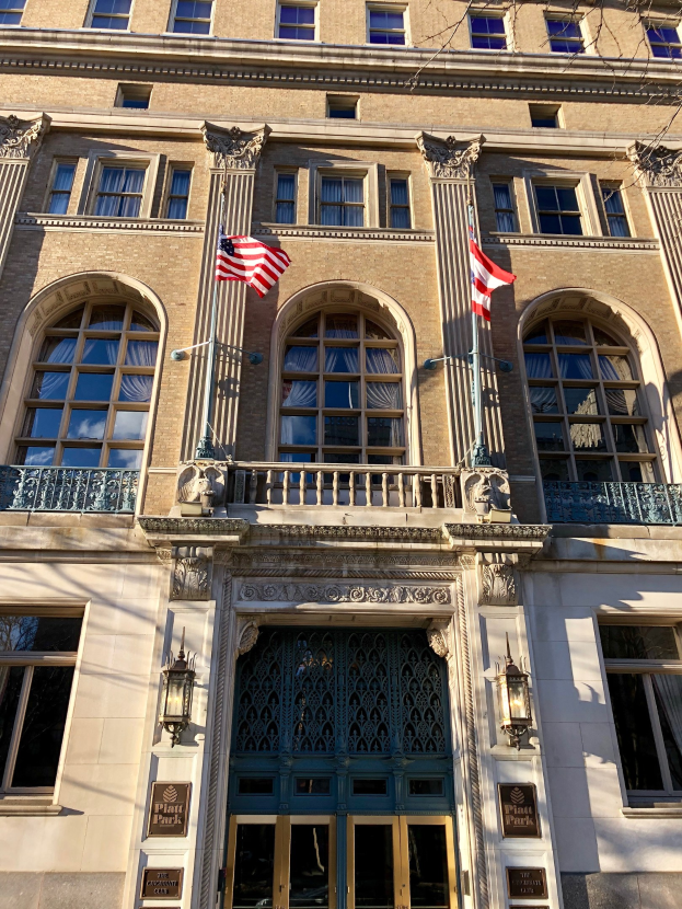 Außenansicht des Plaza Hotels in New York City mit Glasfenstern, -türen, -säulen, -geländern, -leuchten, Texttafeln und Flaggen vor einem Himmelhintergrund.