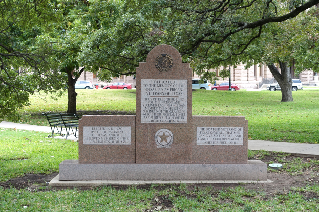 Ein Denkmal steht in einem Park mit einer Bank davor, umgeben von Gras und Bäumen, mit Fahrzeugen, Gebäuden und einem klaren blauen Himmel im Hintergrund.