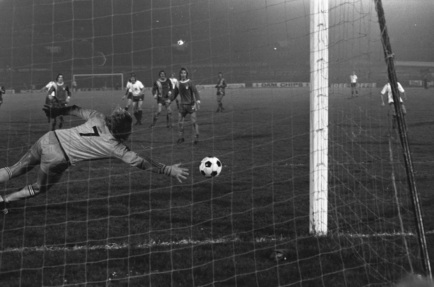Ein Schwarz-Weiß-Foto eines Fußballspielers bei einer Parade vor einem Tor, umgeben von einer Gruppe von Menschen auf dem Boden, Torpfosten sichtbar auf der rechten Seite und Werbetafeln und Lichter im Hintergrund.