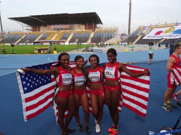 Vier Frauen in Sportkleidung halten amerikanische Flaggen und feiern als Teil eines 4x400m-Staffel-Sieges, umgeben von Zuschauern und Stadion-Elementen.