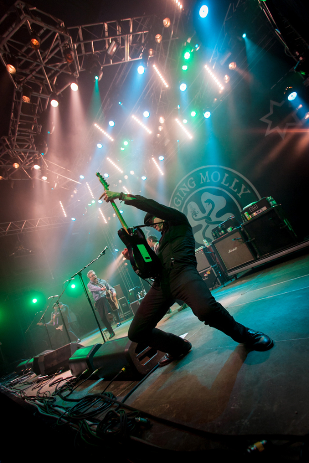 Ein musikalisches Konzert mit drei Performern auf der Bühne: zwei Gitarristen rechts mit Mikrofonen davor, ein Hintergrund mit "MOLLEY" und zwei Schlangenlogos sowie Scheinwerfer darüber.