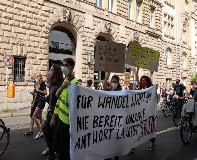 Eine Gruppe von Menschen, die auf einer Demonstration durch die Straße marschieren, einige halten Schilder und andere fahren Fahrräder, mit einem Gebäude im Hintergrund, das Fenster, Bögen, Säulen und Skulpturen, Bäume und einen Lichtmast zeigt.