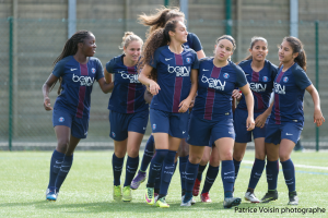 Eine Gruppe junger Frauen beim Fußballspielen auf einem Rasenfeld mit einem Maschendrahtzaun und Pfosten im Hintergrund und einer Wand dahinter, mit dem Text "Paris Saint-Germain Frauenfußball" in der rechten unteren Ecke.