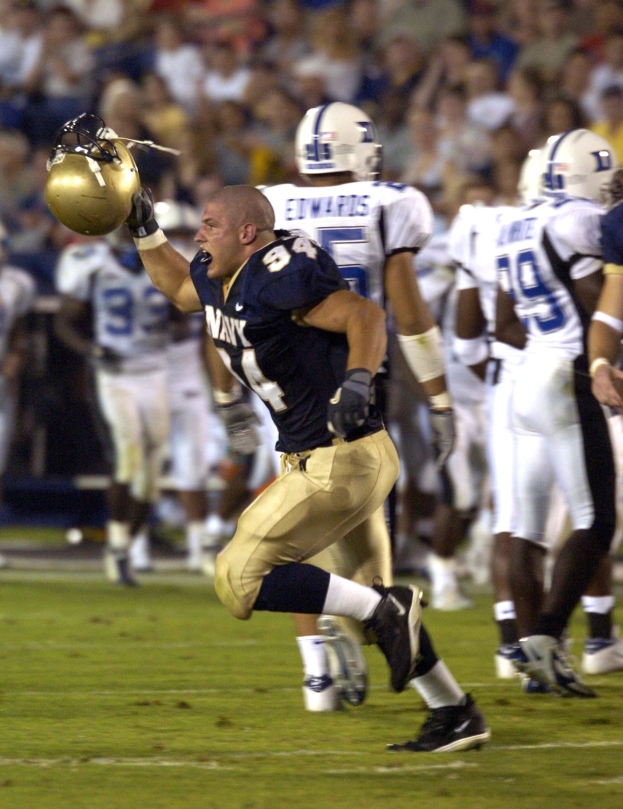Ein Fußballspieler in weißer Uniform rennt mit dem Ball, umgeben von Teamkollegen und Gegenspielern in Helmen und Sportbekleidung auf dem Feld, während Zuschauer im Hintergrund sitzen.