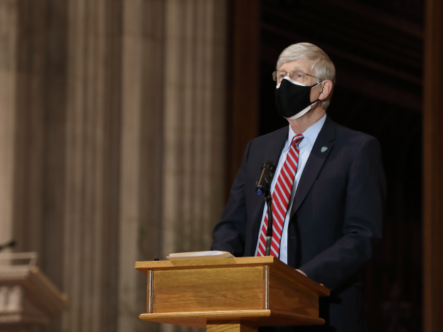 Ein Mann in Anzug und Krawatte mit einer Gesichtsmaske steht an einem Rednerpult mit einem Mikrofon und einem Buch, hält eine Rede mit Säulen und einer Wand dahinter und einem weiteren Rednerpult links von ihm.