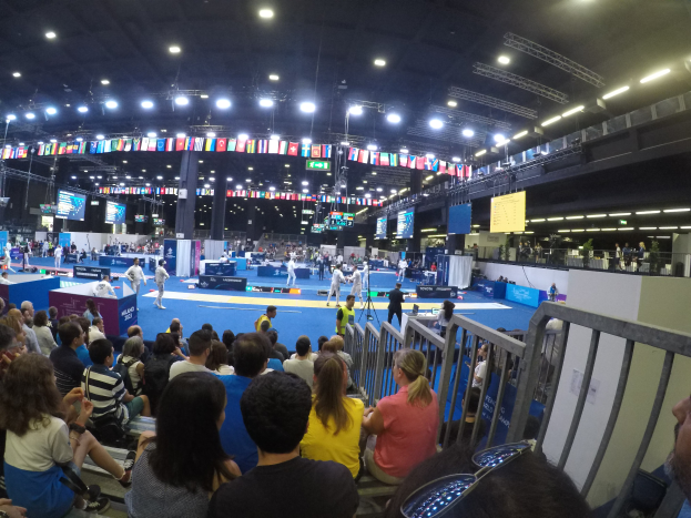 Eine große Menschenmenge, die auf Sitzplätzen und Stehplätzen in einer Halle sitzt, mit einem Geländer auf der rechten Seite, Bildschirmen, Fahnen und Deckenleuchten im Hintergrund.