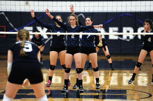 Drei Mädels bei einem Volleyballspiel mit erhobenen Händen, ein Netz davor und ein Zaun im Hintergrund.