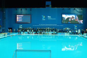 Gruppe von Menschen, die um ein Hallenbad mit Netz am Boden stehen, mit einer Wand im Hintergrund, die den Text und Logos 'FINA World Championships 2015 Kazan' anzeigt, sowie Träger und Lampen.