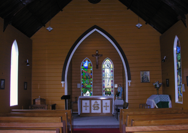 Das Innere einer Kirche mit Bänken auf beiden Seiten, einem Kreuz in der Mitte, hängenden Lampen oben, einem Tisch mit einer Vase und einem gerahmten Bild auf der rechten Seite und einer Statue mit einem Tisch und einem Tuch daneben in der Mitte.