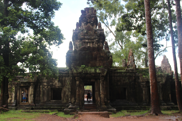 Eine Gruppe von Menschen vor dem Angkor Thom-Tempel in Kambodscha, umgeben von grünem Gras und hohen Bäumen, mit einem sichtbaren Himmel im Hintergrund.