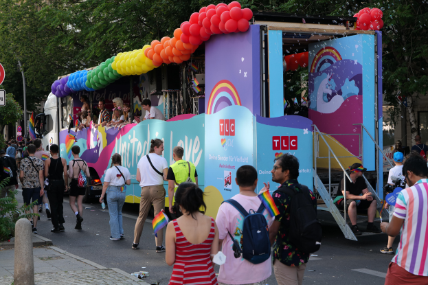 Eine Gruppe von Menschen geht neben einem Lastwagen mit bunten Luftballons die Straße entlang, mit Schildern an Pfählen am Straßenrand und Bäumen und Gebäuden im Hintergrund, was auf eine Pride-Parade in Paris hindeutet.