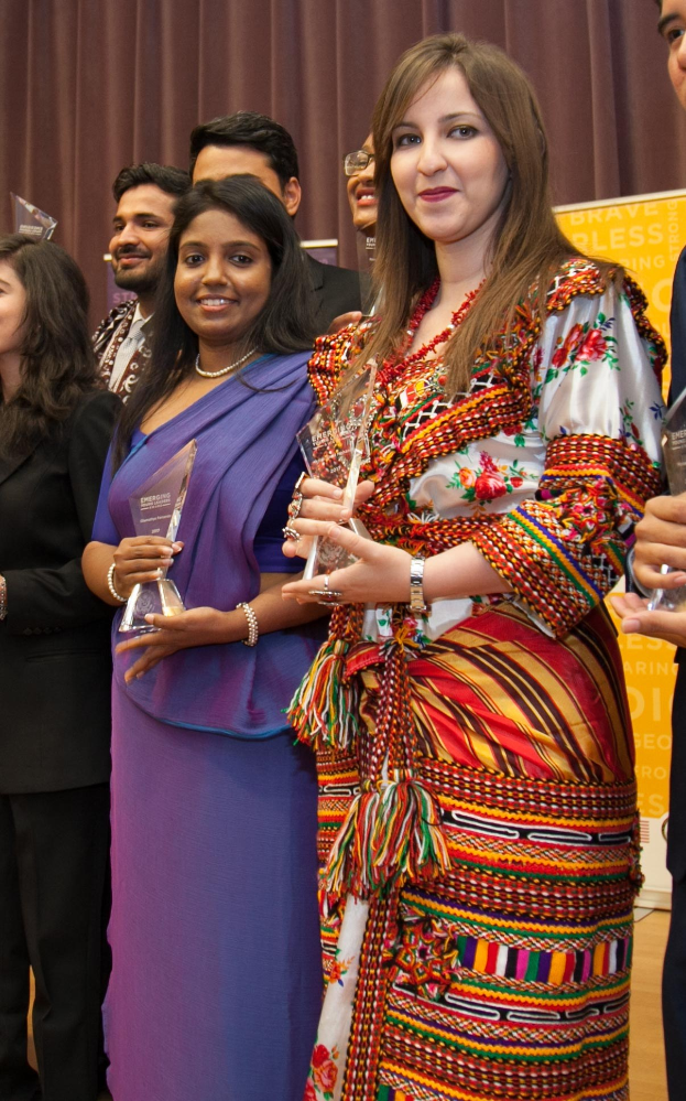 Gruppe von Menschen auf einer Bühne mit einer Frau in der Mitte, die eine Trophäe hält, dahinter ein Vorhang und eine Tafel mit der Aufschrift "Bravo Blessing Award Gewinner."