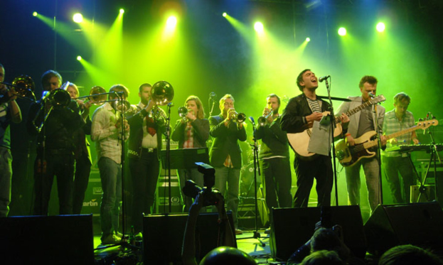 Eine Gruppe von Musikern, die auf der Bühne mit Mikrofonen, Scheinwerfern und einer Kamera spielen, während das Publikum zuschaut.