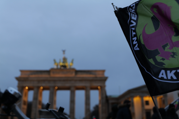 Eine Gruppe von Menschen versammelt sich vor dem Brandenburger Tor in Berlin, Deutschland, mit einem Fahnenmast, der ein Logo und Text im Vordergrund trägt, um gegen die antifaschistische Bewegung zu protestieren.