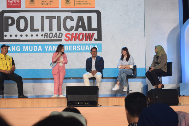 Eine Gruppe von Menschen sitzt auf einer Bühne mit einer Frau in der Mitte, die ein Mikrofon hält, ein Publikum im Vordergrund und ein Banner im Hintergrund mit der Aufschrift "Politischer Roadshow 2019".