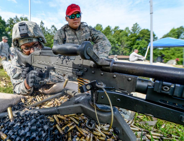 Eine Person auf einem Armee-Trainingsgelände feuert eine Waffe ab, neben ihr steht eine Person mit roter Mütze, während im Hintergrund Bäume, Zelte und Menschen zu sehen sind, an einem klaren, sonnigen Tag, mit Patronenhülsen und Gras im Vordergrund.