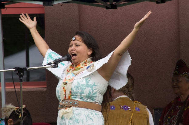 Eine Frau in einem weißen Kleid steht auf einer Bühne, spricht in ein Mikrofon und hat eine Wand und einen Eisenrahmen hinter sich.
