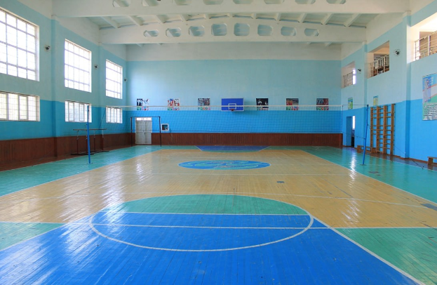 Großer Turnsaal mit einem blauen und weißen Boden, Basketballfeldmarkierungen, einem Netz an Stangen, einer Tür, Fenstern, Wandplakaten, einem Geländer und einer sichtbaren Dachkonstruktion.