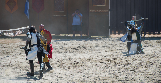 Eine Person steht in der Mitte und hält eine Kamera, während sie ein Bild von einem Kampf zwischen zwei Gruppen auf der rechten und linken Seite aufnimmt, mit einer Wand, einem Tor und einer Fahne im Hintergrund und Sand am Boden.