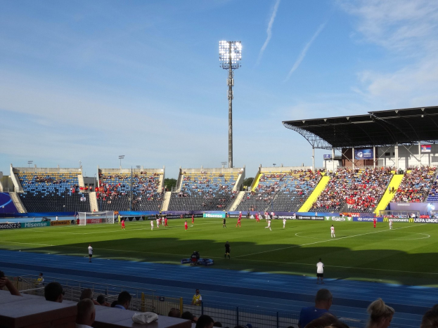 Ein Fußballspiel in einem großen Stadion mit Zuschauern auf den Rängen und Spielern auf dem Feld unter Flutlicht, mit dem Himmel im Hintergrund.