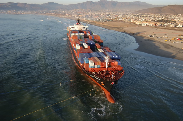 Ein großes Containerschiff fährt auf dem Meer in der Nähe eines Strandes, mit zahlreichen Containern an Bord, während Fahrzeuge am Strand und Gebäude, Bäume, Berge und der Himmel im Hintergrund zu sehen sind.