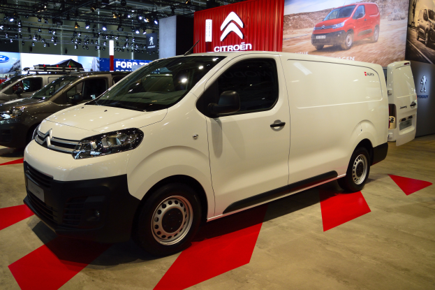 Ein weißer Citroën Berlingo Van wird auf einer Automesse präsentiert und von schwarzen Schlafzimmermöbeln umgeben. Die Wände sind mit Brettern und Lichtern geschmückt.