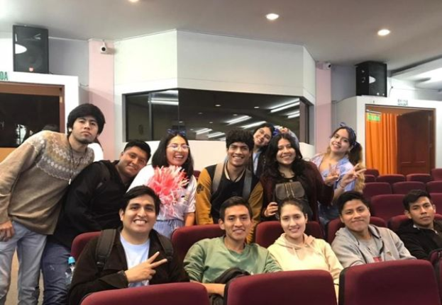 Eine Gruppe von Menschen, die in einem Kino für ein Foto posieren, einige sitzen und einige stehen, alle lachen, mit Lautsprechern an der Wand und Lampen an der Decke im Hintergrund.