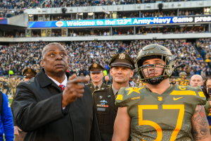 A man in a suit and tie stands next to a football player in a helmet, surrounded by people in uniforms and caps, with a stadium filled with spectators and a board reading "Oregon Football Coach Mike McCarthy and Oregon QB Marcus Mariota" in the background.
