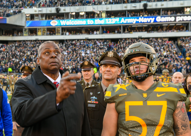 A man in a suit and tie stands next to a football player in a helmet, surrounded by people in uniforms and caps, with a stadium filled with spectators and a board reading "Oregon Football Coach Mike McCarthy and Oregon QB Marcus Mariota" in the background.