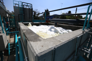 Ein großer Tank voller weißer Schaum steht auf einer Zementwand, umgeben von Metallstangen, Treppen, Geländern und anderen Gegenständen, mit Bäumen, Pfählen, Drähten und einem klaren blauen Himmel im Hintergrund.