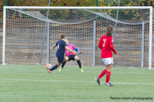 Eine Gruppe von Frauen, die auf einem grünen Rasenfeld Fußball spielen, umgeben von Bäumen, mit einem Tor im Hintergrund und Text am unteren Rand des Bildes.