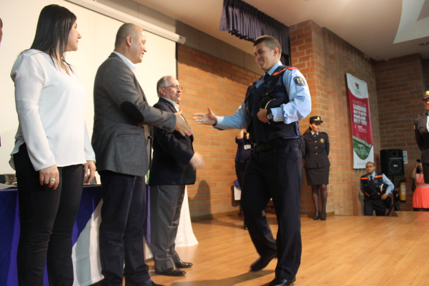 Eine Gruppe von Menschen auf einer B√ľhne, einer in Uniform mit einer M√ľtze, eine Frau in einem wei√üem Hemd, mit einem Banner, Lautsprecher, Vorh√§ngen, Tisch und Wandbildschirm im Hintergrund.