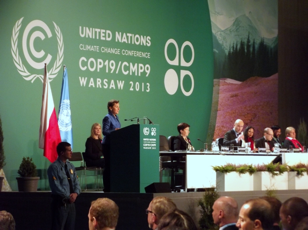 Eine Frau steht neben einem Podium mit Mikrofonen und spricht zu einer sitzenden Menge; im Vordergrund befindet sich ein Tisch mit Gläsern, Mikrofonen, Namensschildern und anderen Gegenständen sowie zwei Fahnen an Ständern und ein Blumentopf mit einer Pflanze daneben, während im Bild unten eine weitere Person und eine Gruppe von Menschen zu sehen sind.