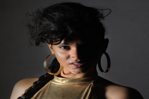 Eine Frau in einem goldenen Kleid und großen Ohrringen steht selbstbewusst vor einer Wand, ihr glatter Bob-Haarschnitt und das natürliche Make-up unterstreichen ihren entschlossenen Gesichtsausdruck, während sie direkt in die Kamera schaut.