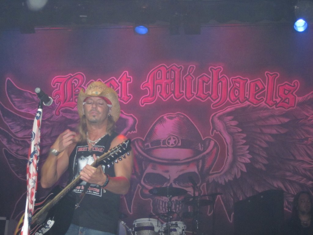 Eine Musik-Konzertszene mit Scheinwerfern oben, einem Banner im Hintergrund, Drogen in der Mitte unten, einer Person in der rechten unteren Ecke sitzend und einem Gitarristen mit Mikrofon links.