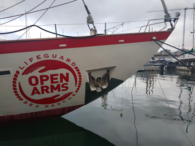 Ein rotes und weißes Boot mit der Aufschrift "Open Arms Sea Rescue" liegt im Wasser mit mehreren anderen Booten, Pfählen und einem klaren blauen Himmel im Hintergrund.