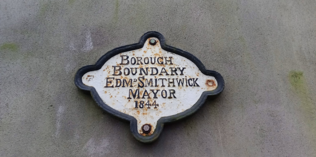 Metallplatte an der Seite eines Gebäudes, die die Borough-Grenze markiert, mit schwarzem Hintergrund und weißer, fettgedruckter, zentrierter Schrift, die "Borough Boundary Edm Smithwick Mayor" lautet.