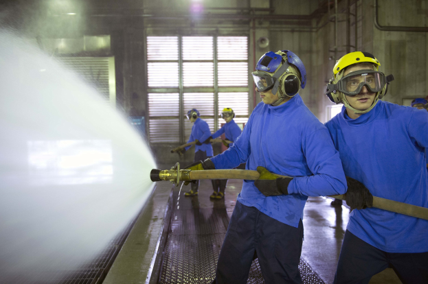 Gruppe von Männern in blauen Hemden und gelben Helmen bei der Arbeit an einer Maschine in einer Fabrik, wobei einer das Boden mit Wasser besprüht.