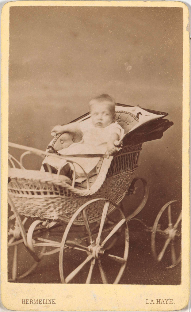 Ein altes Schwarz-Weiß-Foto eines Babys in einem hölzernen Kinderwagen mit vier Rädern, das ein weißes Strampler trägt und einen friedlichen Ausdruck hat, mit kursiver Schrift unten.