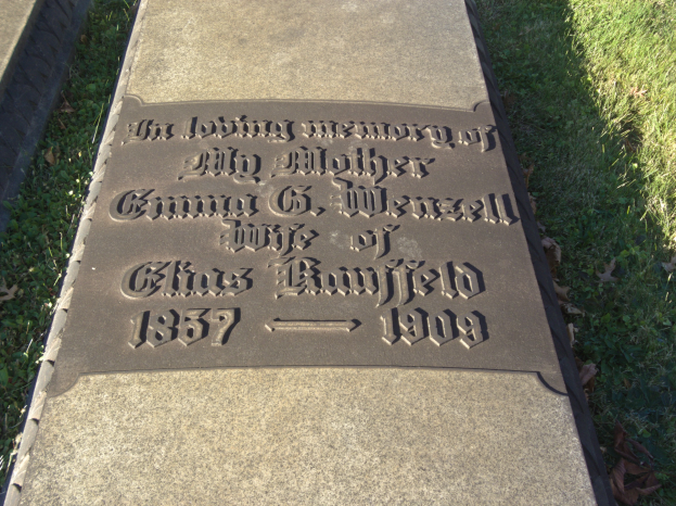 Grabstein auf einem Friedhof mit der Inschrift "Die liebevolle Erinnerung an meine Mutter", umgeben von Gras und trockenen Blättern.