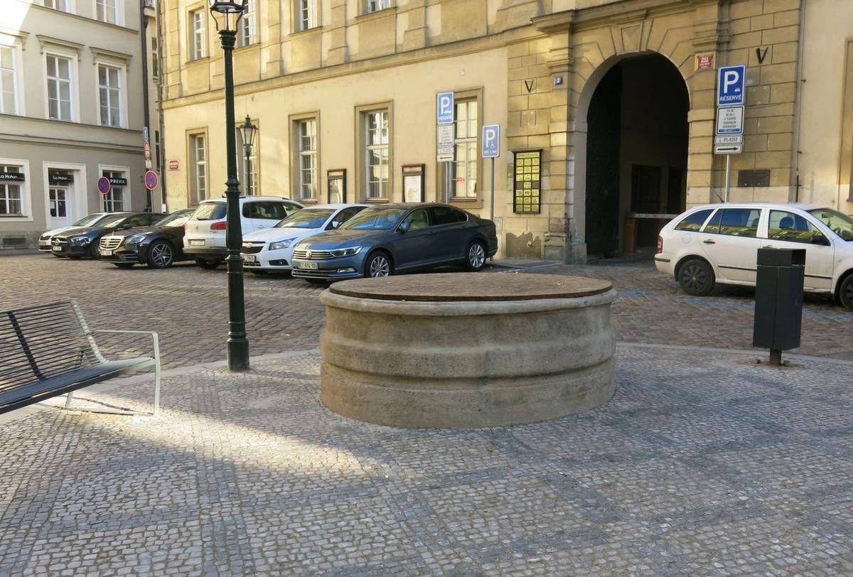 Ein Stadtplatz mit einer zentralen Bank, einem Brunnen, Laternen und Gebäuden mit Fenstern und Schildern.