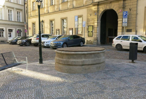 Ein Stadtplatz mit einer zentralen Bank, einem Brunnen, Laternen und Gebäuden mit Fenstern und Schildern.