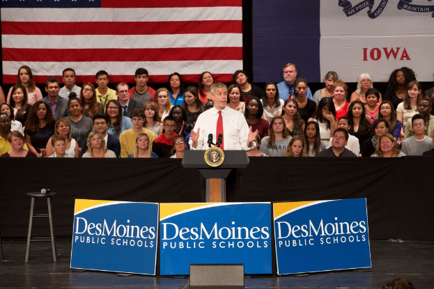 Ein Mann in einem weißen Hemd und roter Krawatte steht an einem Rednerpult mit drei Tafeln, einem Hocker mit Mikrofon und einer sitzenden Menge im Hintergrund, sowie einer Flagge und zusätzlichem Text.
