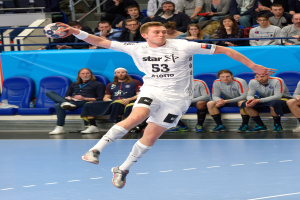 Ein Mann in der Luft, der einen Fußball hält, umgeben von sitzenden Menschen und einem Geländer im Hintergrund, trägt Schuhe.