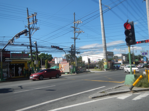Ein rotes Auto fährt auf einer Straße neben einer Ampel, mit Strommasten und Drähten entlang der Straße, Gebäuden und Bäumen im Hintergrund und einem klaren blauen Himmel.
