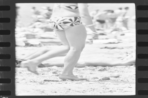 Eine Frau in einem Bikini geht am Strand spazieren, im Hintergrund liegen ein paar Menschen auf dem Boden, dargestellt in Schwarz-Weiß.