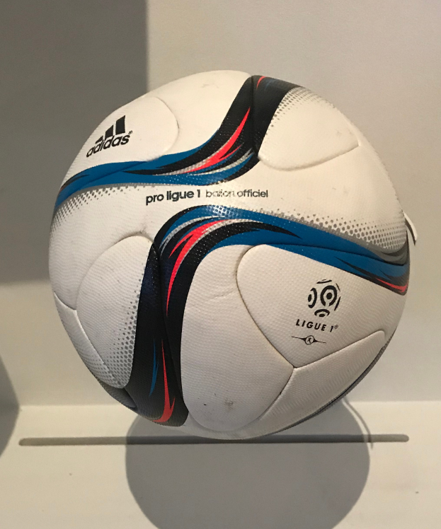 Ein weißer Fußball mit dem Adidas-Logo und "Ligue 1" in fetter schwarzer Schrift auf einem weißen Tisch gegen einen hellen Hintergrund.
