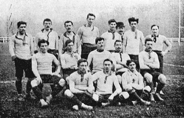 Ein Schwarz-Weiß-Foto einer Fußballmannschaft aus den 1920er Jahren, mit einigen Spielern stehend und anderen auf dem Boden sitzend, vor einer Kulisse aus Pfählen, Bäumen und einem klaren Himmel.
