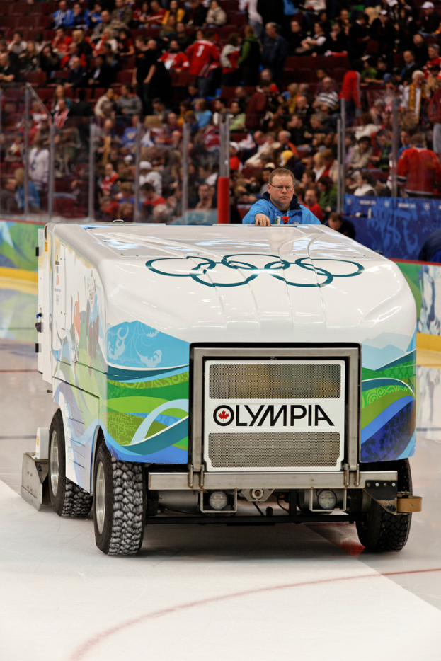 Ein Mann in einem blauen Jackett und Brille steht auf einem Fahrzeug, mit einem Publikum auf Stühlen darüber und einem Zaun und Werbetafel im Hintergrund.