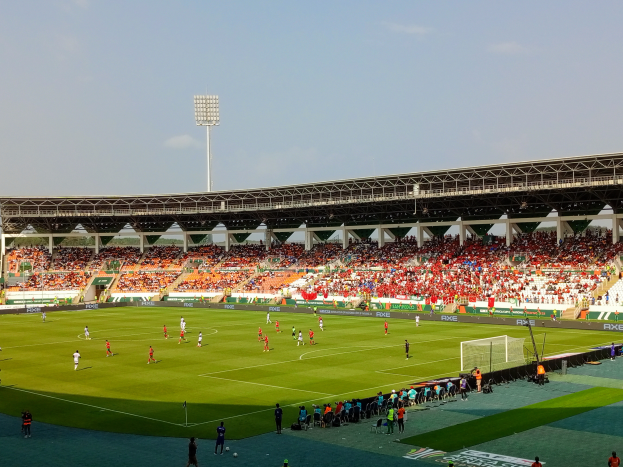 Ein Fußballspiel wird in einem großen Stadion mit Menschen auf dem Boden, einem Torpfosten, einem Zaun, einer Fahne mit Text, Metallrahmen, einem Dach mit Deckenleuchten und einem bewölkten Himmel gespielt.